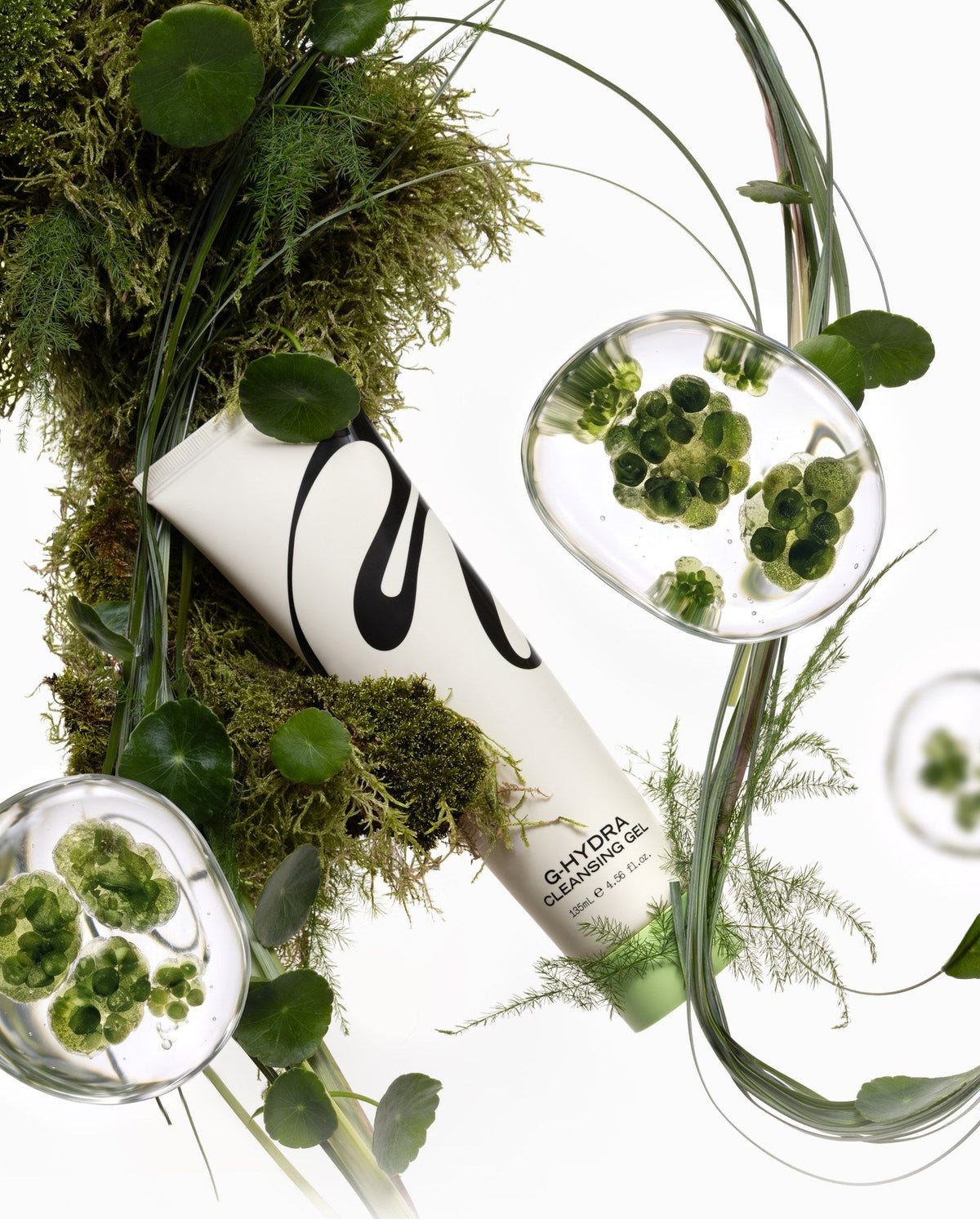 A white cosmetic tube with black script branding lies diagonally on a white background, surrounded by green moss, leaves, and clear oval glass droplets containing green botanical elements.